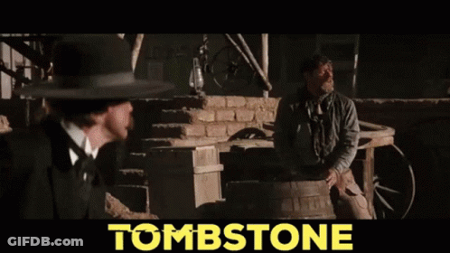Man In Cowboy Hat Holding Gun In Front Of Tombstone GIF