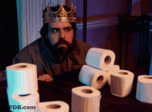 Man In Crown Sits At Table With Toilet Paper Rolls GIF