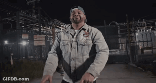 Man In Denim Jacket And Hat Standing In Front Of Fence GIF