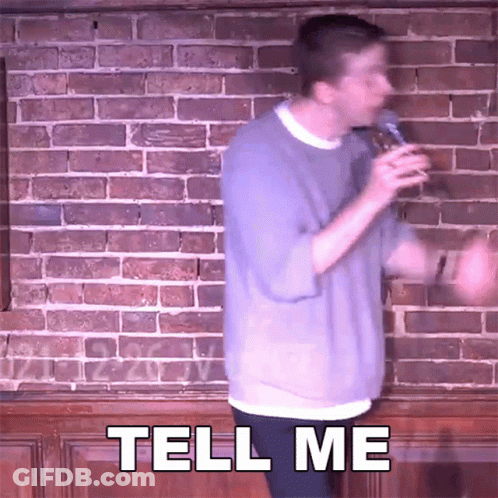 Man In Front Of Brick Wall Holding Microphone Says Tell Me GIF