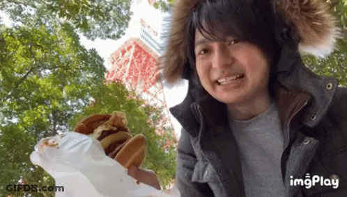 Man In Fur Hood Holding Hamburger In Front Of Tower And Trees GIF