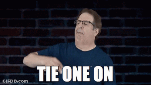 Man In Glasses And Blue Shirt Says Tie One On In Front Of Brick Wall GIF