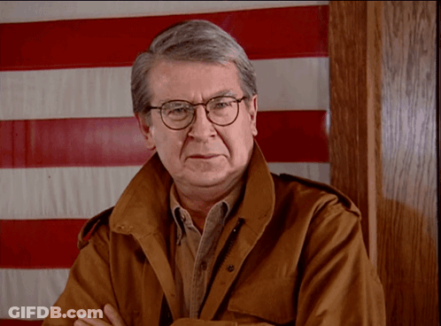 Man In Glasses And Brown Jacket In Front Of Red And White Striped Flag GIF