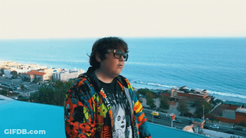 Man In Glasses And Colorful Jacket On Balcony Overlooking City And Ocean GIF