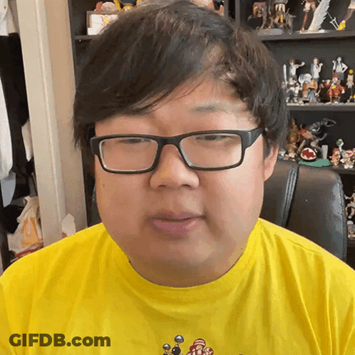 Man In Glasses And Yellow Shirt Sitting In Front Of Toy Shelf GIF