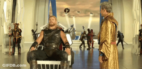 Man In Gold Robe Standing Next To Bearded Man In Chair GIF