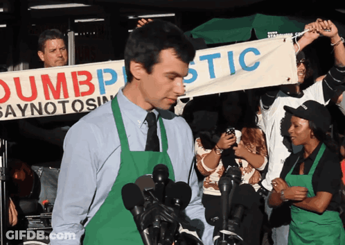 Man In Green Apron Holding Dumbplastic Banner GIF