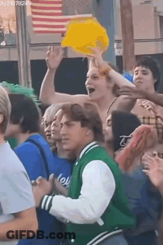 Man In Green Jacket Holding Yellow Hat In Crowd GIF