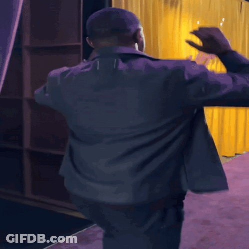 Man In Grey Jacket Dancing On Purple Carpet In Front Of Yellow Curtain GIF