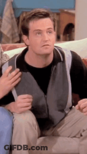 Man In Grey Vest Sitting On Couch Talking To Another Man GIF
