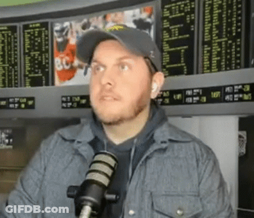 Man In Hat And Jacket Talking Into Microphone At Betting Board GIF