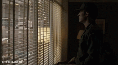 Man In Hat Looking Out Window With Blinds GIF
