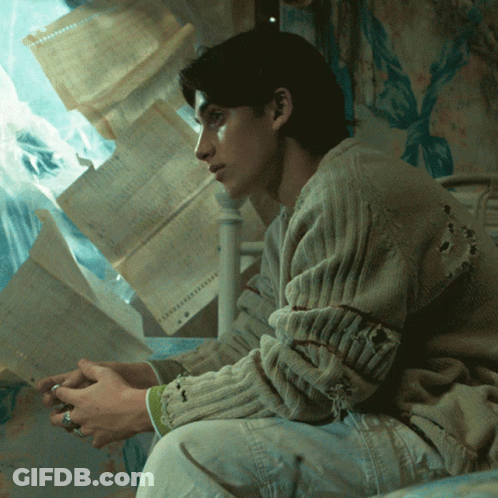 Man In Hole Sweater Sitting On Bed With Papers GIF