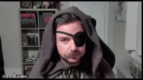 Man In Hooded Cape And Eye Patch Sitting In Front Of Bookshelf GIF