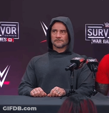 Man In Hoodie Sitting At Table With Microphones In Front Of Surv Sign GIF