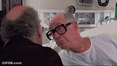 Man In Hospital Gown Looking At Man With Glasses GIF