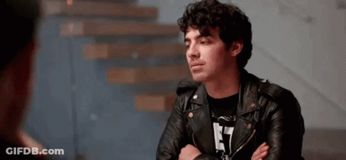 Man In Leather Jacket Sitting With Arms Crossed Talking To Another Man GIF