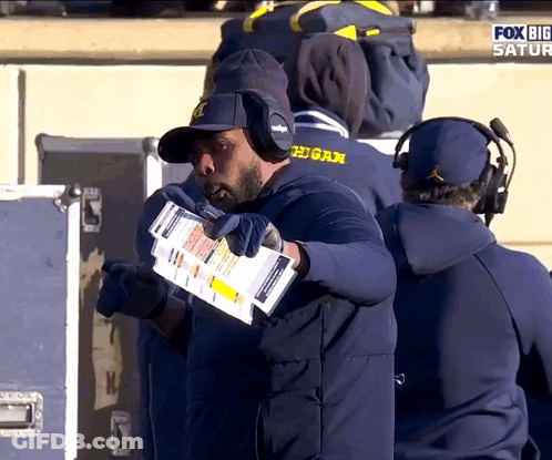 Man In Michigan Hat Holding Paper In Mouth GIF