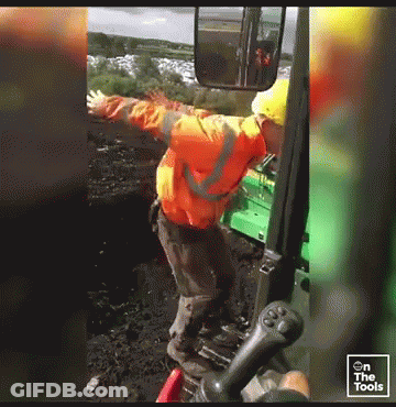 Man In Orange Jacket And Yellow Hard Hat In Front Of Green Tractor With Arms Outstretched GIF