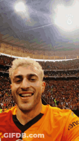 Man In Orange Shirt Saying Eme Smiling In Crowd GIF