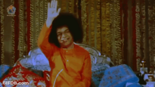 Man In Orange Shirt Sitting On Bed Waving GIF