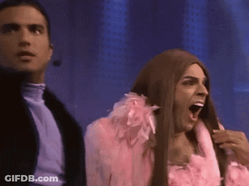 Man In Pink Feathered Coat Next To Woman In Wig GIF