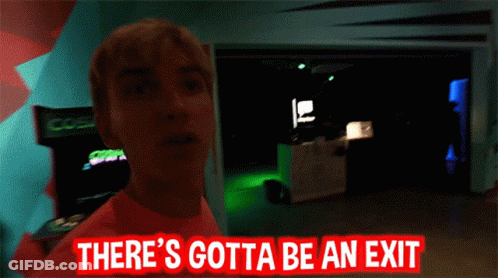 Man In Pink Shirt Standing In Dark Room With Sign Somewhere Over Here GIF