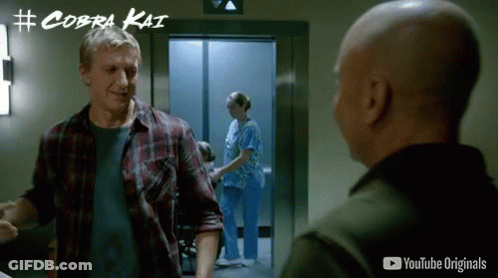 Man In Plaid Shirt Talking To Another Man In Front Of Elevator With Cobra Kai Written On Wall GIF