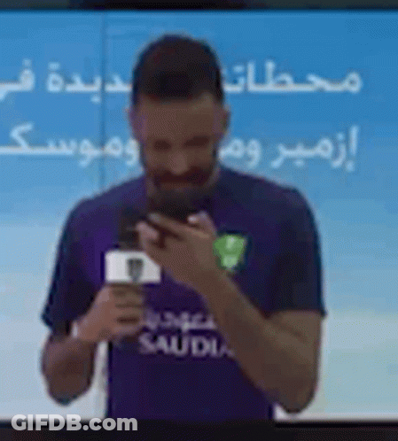 Man In Purple Shirt Holding Phone In Front Of TV GIF