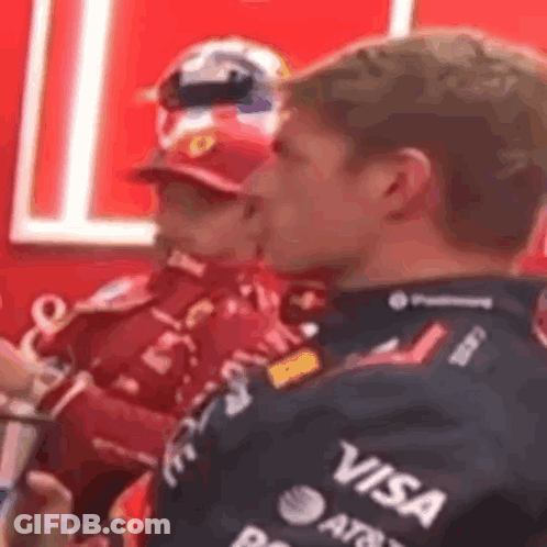 Man In Red And Black Jacket With Visa Sitting Next To Another Man GIF