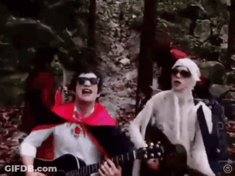 Man In Red Cape Playing Guitar Next To Man GIF