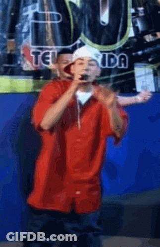 Man In Red Shirt Singing Into Microphone In Front Of Teiuda Sign GIF