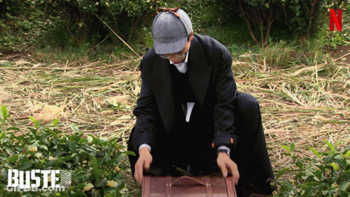 Man In Suit And Hat Kneeling With Suitcase In Front Of Buste Sign GIF