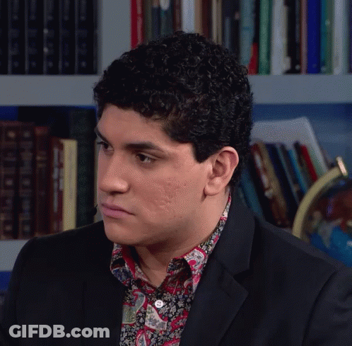 Man In Suit And Paisley Shirt Sitting Before Bookshelf GIF