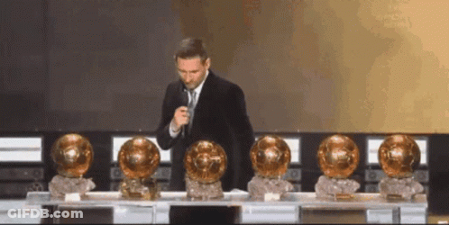 Man In Suit And Tie At Microphone With Gold Trophies GIF