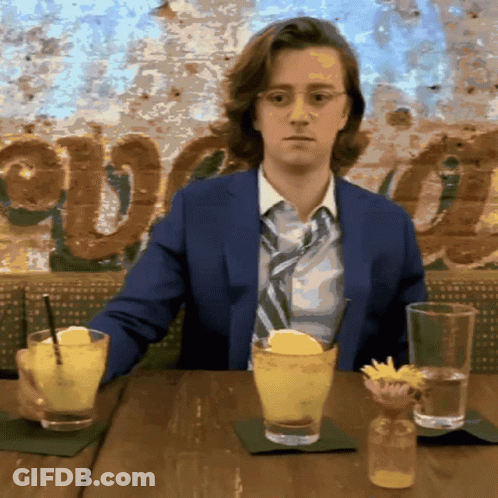 Man In Suit And Tie At Table With Drinks And Avocado Sign GIF