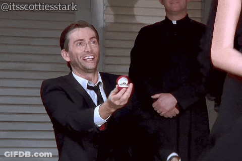 Man In Suit And Tie Holding Engagement Ring To Woman GIF