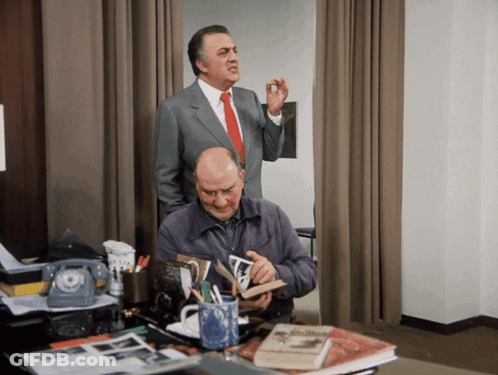 Man In Suit Behind Man At Desk With Book GIF