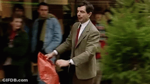 Man In Suit Holding Red Bag Next To Christmas Tree GIF