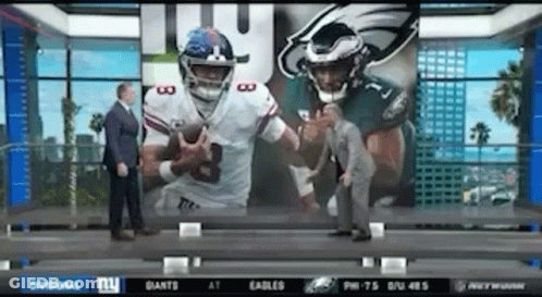 Man In Suit In Front Of Screen With Two Football Players GIF