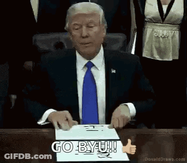 Man In Suit Sitting At Table With Go BYU Sign GIF