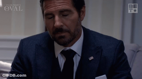 Man In Suit Sitting Before Oval Sign GIF