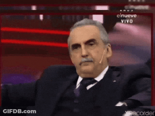 Man In Suit Sitting Before Screen Saying El Nueve Vivo GIF