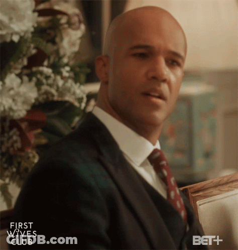 Man In Suit Sitting In Front Of First Wives Club Sign GIF