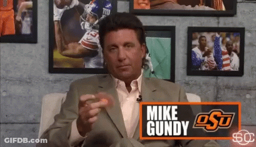 Man In Suit Sitting In Front Of Mike Gundy Sign GIF