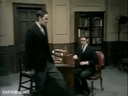 Man In Suit Standing Beside Man In Chair GIF