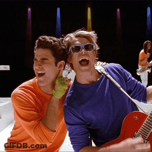 Man In Sunglasses Singing Into Microphone Beside Man In Orange Shirt GIF
