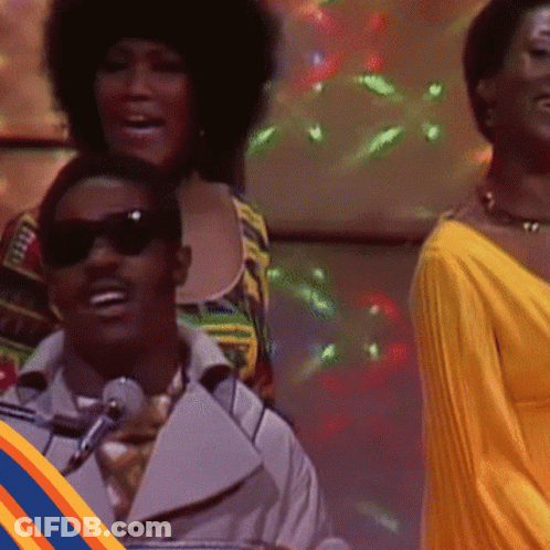 Man In Sunglasses Singing Into Microphone Woman In Yellow Dress Behind GIF