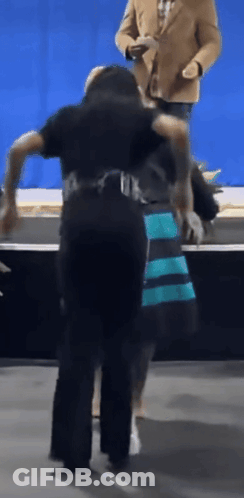 Man In Tan Jacket Behind Woman In Blue And Black Dress GIF