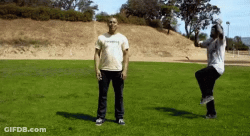 Man In Thrasher Shirt Jumping While Another Man Stands In Grass GIF